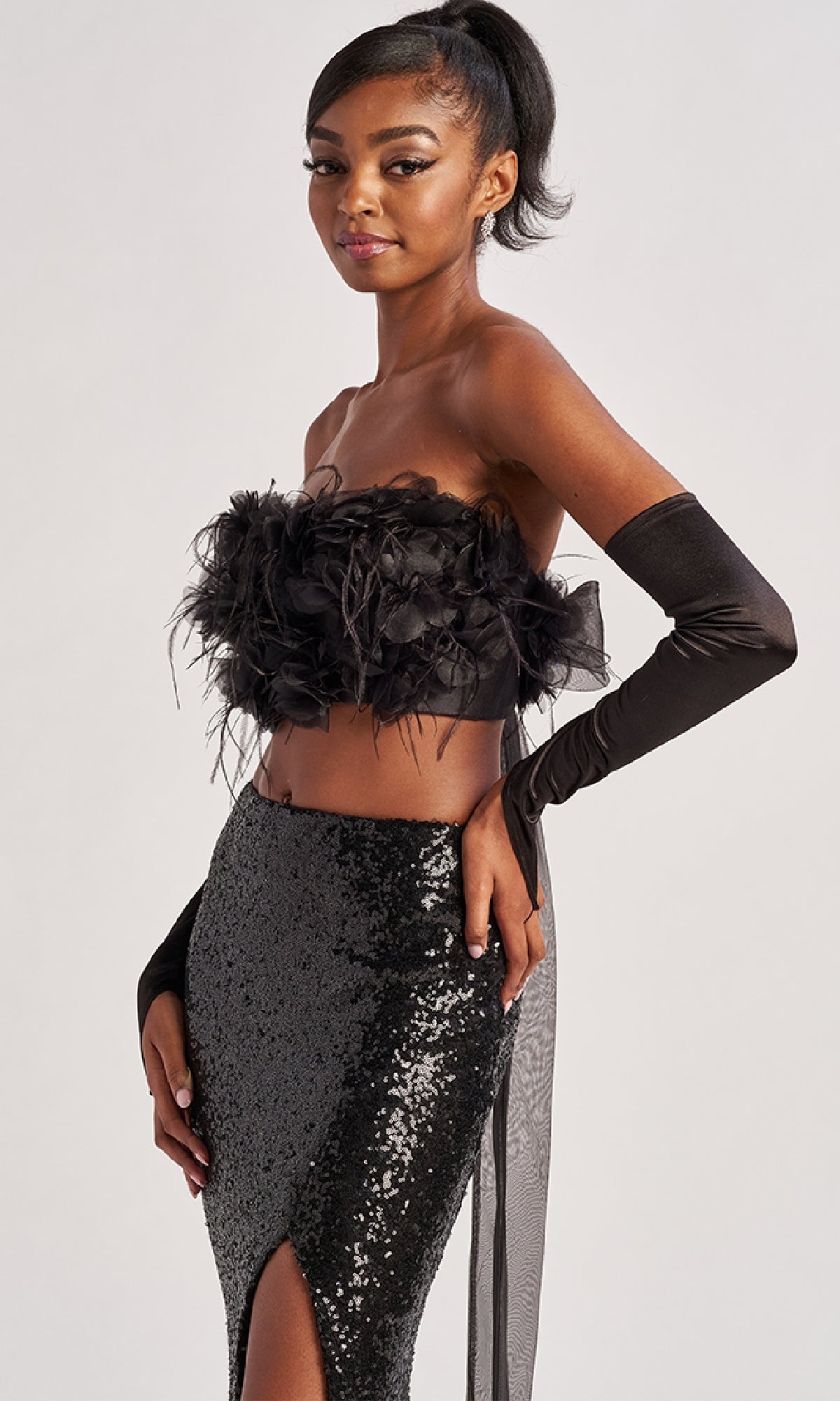 A woman exudes confidence in the Colette CL8450 long prom dress, featuring a feathered crop top and sequined floor-length skirt with a thigh-high slit, paired with elegant black gloves against a plain backdrop.